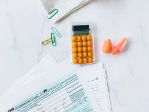 Documentos fiscales, clips de colores y una calculadora naranja sobre una mesa blanca, representando el proceso de cálculo de costes y presupuesto necesario para comprar una casa