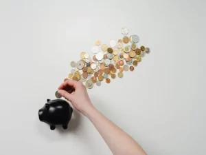 Mano de una persona insertando una moneda en una hucha negra, con muchas monedas esparcidas en el fondo blanco, simbolizando el ahorro para la compra de una casa