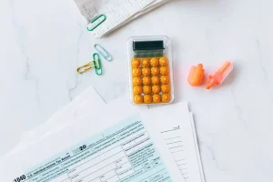 Tax documents, colorful paper clips, and an orange calculator on a white table, representing the process of calculating costs and budget needed for buying a home