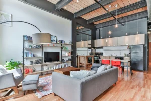 Espai d’estil loft amb disseny industrial, que inclou una sala d’estar amb sofà gris, prestatgeries plenes de llibres, llum d’arc, catifa decorativa i una cuina oberta amb mobles de fusta clara, electrodomèstics negres i làmpades penjants.