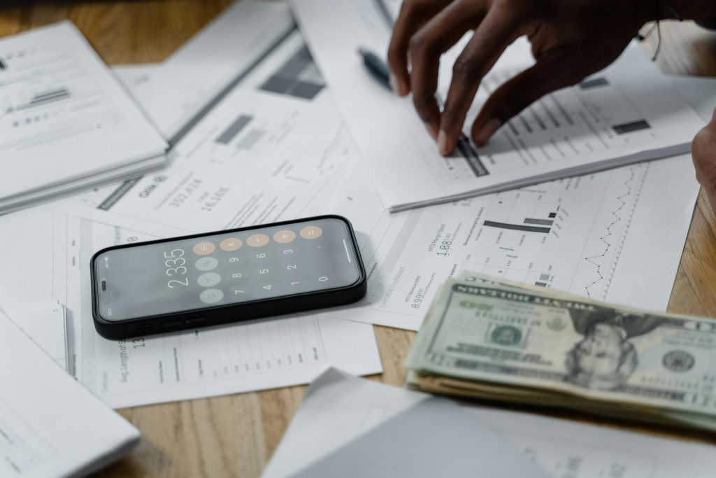 Una persona revisando documentos financieros con una calculadora en su teléfono móvil y una pila de billetes en la mesa, ilustrando la gestión de suministros y finanzas en un piso de alquiler.