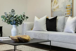Living room with a gray sofa, decorative cushions, and a white vase with green branches on a coffee table, showing how to combine minimalist style with cozy elements to create a balanced and comfortable home environment.