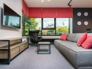 Sala de estar moderna con sofá gris, cojines rojos, una silla negra de diseño y grandes ventanales con persianas rojas, mostrando cómo maximizar el espacio en un salón pequeño con un diseño contemporáneo