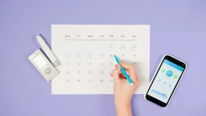 Hand marking dates on a calendar next to a health monitoring device and a mobile phone, symbolizing the importance of organizing and monitoring health effectively.