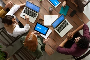 Personas trabajando en equipo alrededor de una mesa con portátiles y tabletas, compartiendo información durante una reunión, representando la importancia de la colaboración para mejorar la productividad en proyectos empresariales.