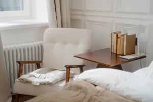 Rincón de lectura en un dormitorio con una silla beige de estilo vintage, mesa auxiliar de madera con libros y una lámpara decorativa.