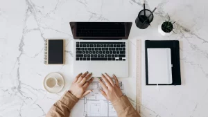 Vista de una mesa de mármol blanca con un ordenador portátil, una taza de café y libretas, con manos sobre el teclado.