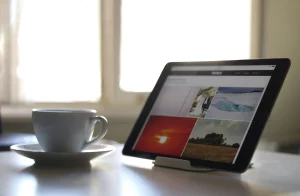 Taza blanca junto a una tablet con imágenes en pantalla sobre una mesa, con luz natural al fondo.