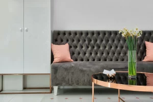 Sala con un sofá de terciopelo gris con diseño capitoné, decorado con cojines rosados. Frente al sofá, una mesa de centro con detalles en metal dorado y un jarrón de cristal con flores blancas completan el estilo sofisticado y minimalista. A la izquierda, un armario blanco con acabados modernos se integra al ambiente.