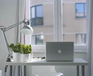  Escritorio blanco con una lámpara de escritorio, dos plantas en macetas blancas, y una MacBook. La imagen muestra una ventana con vista al exterior, con luz natural iluminando el espacio, creando un ambiente relajante y moderno. 