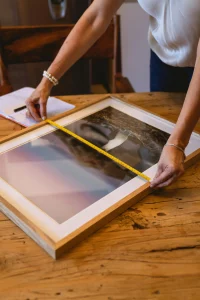 Primer plano de una persona midiendo un cuadro enmarcado con una cinta métrica sobre una mesa rústica de madera. En la escena también hay un cuaderno con notas y un bolígrafo. 