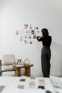 Mujer de espaldas vistiendo ropa elegante y minimalista mientras organiza un moodboard con imágenes de diseño en una pared blanca. En la escena también hay una mesa con revistas y una silla moderna.