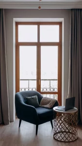 Sillón azul con cojines, mesa de diseño dorada y ordenador portátil junto a una gran ventana. 