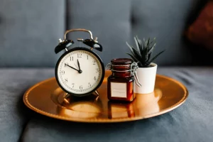 Primer plano de un reloj de alarma clásico negro, un pequeño frasco de vidrio marrón con tapa y una planta en maceta blanca, todos sobre una bandeja dorada. El fondo es de un sofá gris, creando una atmósfera tranquila y elegante. 