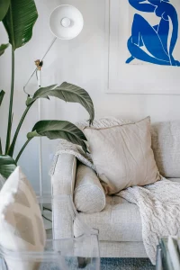 Sala de estar con un sofá beige decorado con cojines y una manta tejida. A su lado, una lámpara blanca de pie, una planta de hojas grandes y un cuadro con una ilustración azul en la pared, creando un ambiente acogedor y elegante.