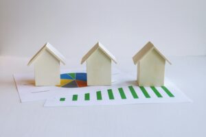 Imagen que muestra tres pequeñas casas de madera sobre gráficos financieros, incluyendo un gráfico de barras en verde que indica crecimiento y un gráfico circular multicolor.