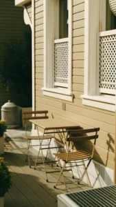 Pequeña terraza urbana con mesa y dos sillas plegables de metal en tonos beige, junto a una fachada de madera clara con celosías decorativas.
