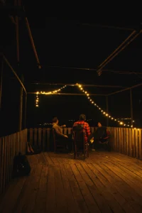 Tres personas conversan en una terraza de madera por la noche, iluminada con guirnaldas de luces cálidas, creando un ambiente íntimo y relajado.