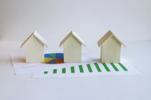 Three small wooden house models placed on top of financial charts, including a pie chart and a bar graph, symbolizing property investment and housing market analysis