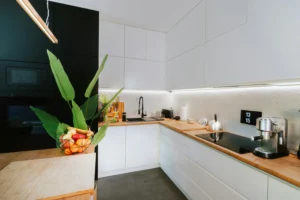 Cocina de diseño contemporáneo con muebles blancos, encimeras de madera, electrodomésticos modernos, iluminación LED y un frutero con hojas verdes que aporta un toque natural.