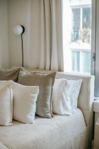 Primer plano de una cama con varios cojines en tonos blanco y beige, colcha texturizada y cortinas claras junto a una ventana luminosa.