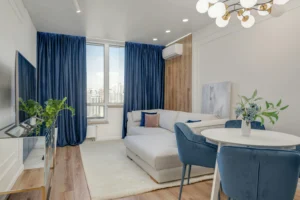Sala de estar luminosa con sofá en forma de L, cortinas azul oscuro, pared de madera, mesa redonda con sillas tapizadas y decoración floral en tonos suaves.