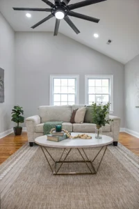 Spacious living room with beige sofa and neutral-tone cushions, round coffee table with books and plants, natural fiber rug and modern ceiling fan