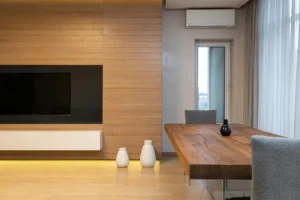 Salón contemporáneo con revestimiento de madera, televisión empotrada, mesa de comedor rústica y sillas grises junto a una ventana con cortinas translúcidas.