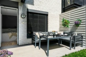 Espacio exterior con suelo de madera clara, sofá en L con cojines, mesa de comedor de cristal y pared decorativa con macetas colgantes, junto a una puerta que conecta con el dormitorio.