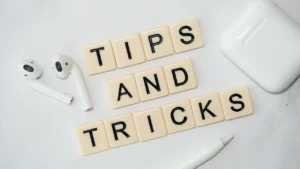 The words "Tips and Tricks" arranged with beige letter tiles, surrounded by wireless earphones, a stylus, and a charging case on a white background