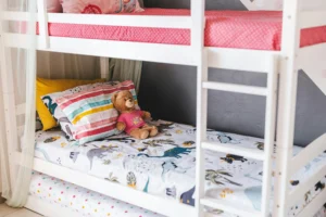 White bunk bed in a child’s bedroom, with colorful bedding, printed cushions, and a teddy bear on the lower bunk