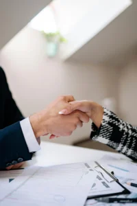 Primer plano de un apretón de manos entre dos personas vestidas formalmente, con documentos y gráficos financieros sobre la mesa