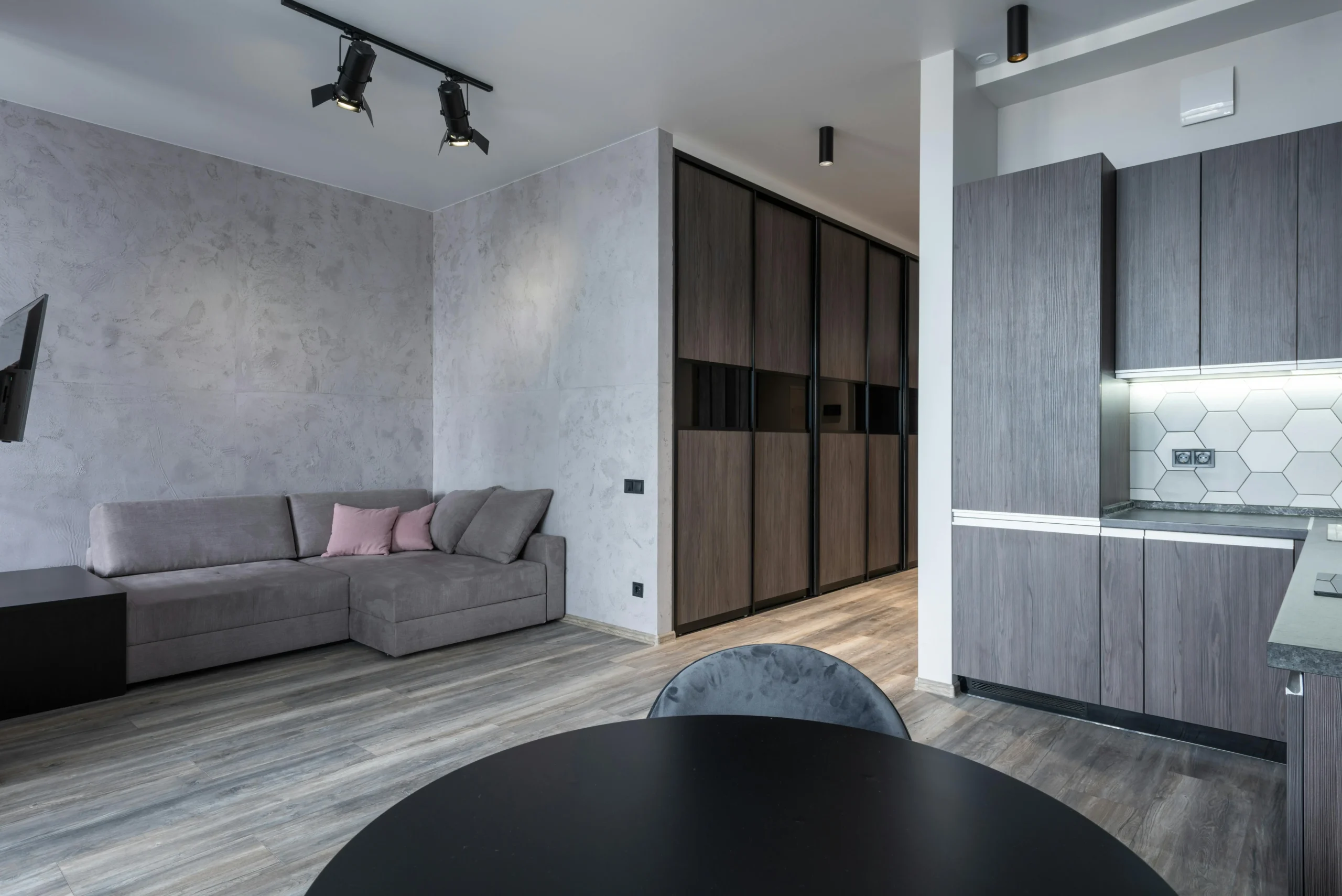 Interior de apartamento con diseño moderno y tonos grises, que incluye un sofá en L con cojines rosa claro, cocina abierta con muebles laminados en gris, salpicadero hexagonal blanco, mesa redonda negra y armarios empotrados en el pasillo