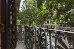 Vista desde un balcón clásico de Barcelona con barandilla de hierro forjado decorativa, vista a una calle arbolada con motos aparcadas y edificios al frente. El sol se filtra entre los árboles creando una atmósfera urbana agradable.