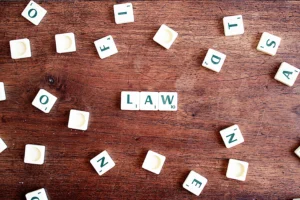 Fichas de juego estilo Scrabble formando la palabra "LAW" en el centro de una mesa de madera, rodeadas de letras sueltas desordenadas