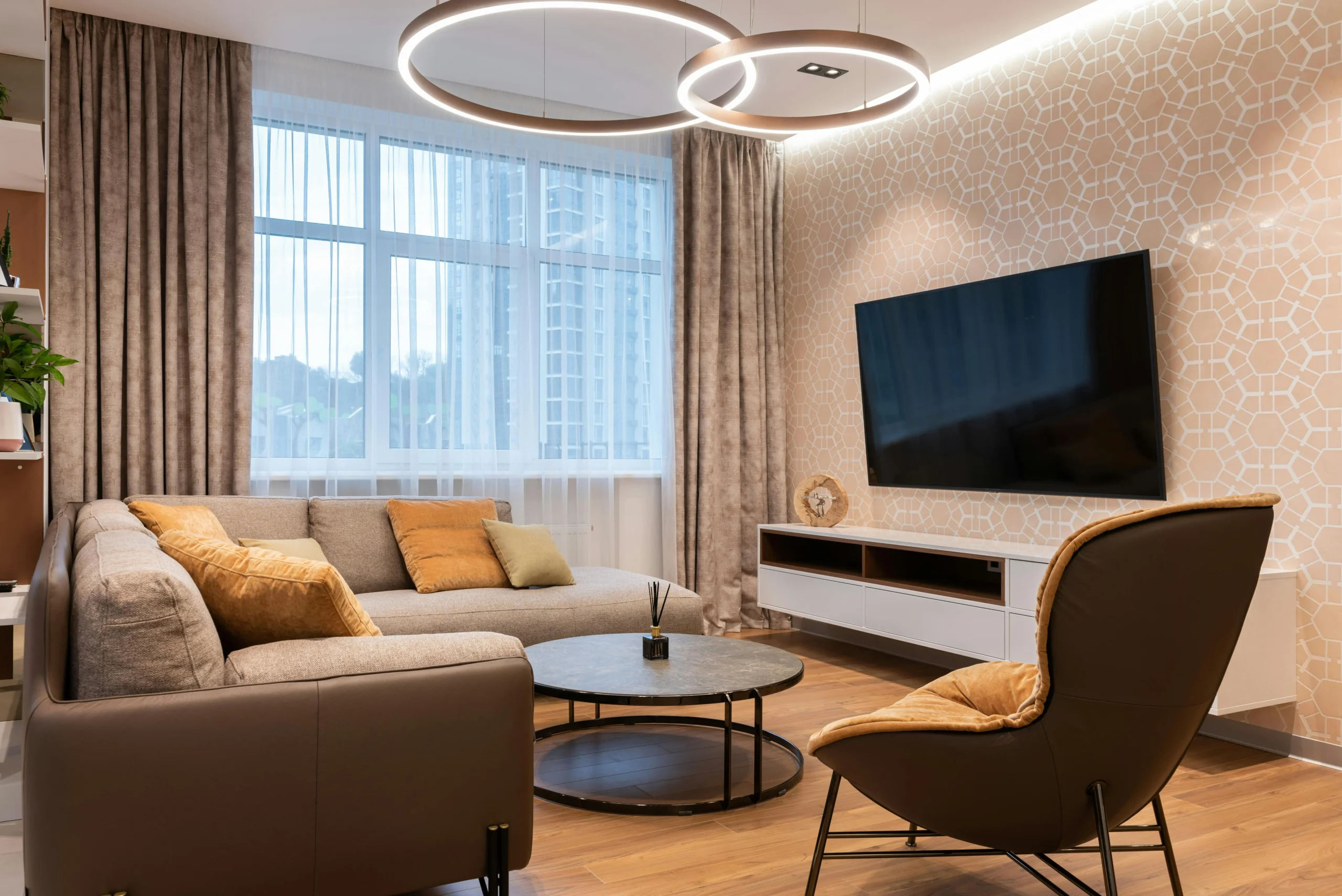 Salón elegante con sofá en L de color beige, cojines mostaza y verdes, mesa de centro redonda negra, televisor de pantalla plana en una pared decorativa y butaca marrón con tapizado suave, bajo lámparas circulares colgantes