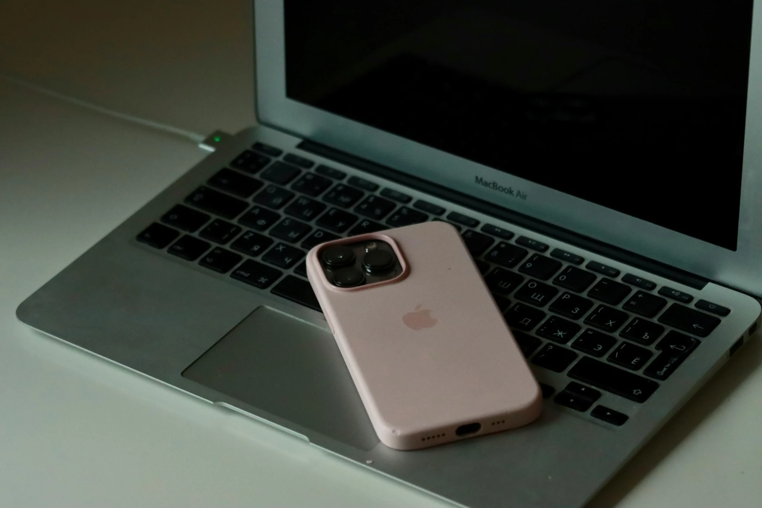 Un iPhone con funda rosa colocado sobre el teclado de un MacBook Air con pantalla apagada, conectado a la corriente, en un entorno de trabajo doméstico