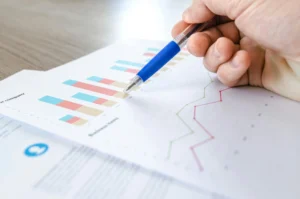 Un primer plano de una mano sosteniendo un bolígrafo azul, señalando gráficos y tablas de datos con barras y líneas, representando un análisis empresarial.