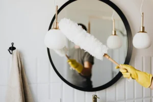 Una persona con guantes amarillos utiliza un plumero blanco para limpiar una lámpara del baño, reflejada en un espejo redondo sobre el lavabo