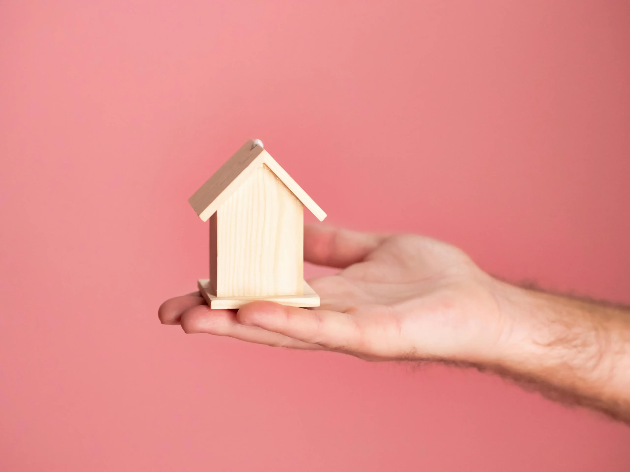 Hand holding a small wooden house model against a pink background