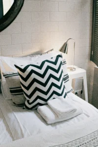 Cama hecha con cojín blanco y negro en zigzag, almohadas rayadas y toallas dobladas. Lámpara metálica en mesita blanca, pared con azulejos, ambiente moderno y acogedor.
