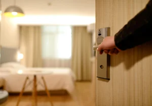 Una persona abre una puerta de madera con cerradura de tarjeta, revelando una habitación de hotel bien iluminada con cama, cortinas y una mesa pequeña. El foco está en la mano y la manija de la puerta, con la habitación desenfocada al fondo.