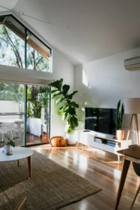 Un salón moderno iluminado por el sol con techos altos, ventanales, alfombra acogedora, mesa blanca, TV en mueble bajo y plantas. Las puertas correderas se abren a un patio con asientos exteriores.