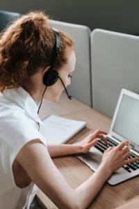 Un agente de soporte al cliente con cabello rizado rojo usando auriculares, escribiendo en una laptop mientras está sentado en un escritorio, enfocado en brindar asistencia.
