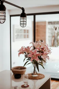 Un jarrón de cristal con lirios rosas frescos sobre un mantel tejido en una isla de cocina blanca. También hay un cuenco de mimbre, una taza negra y un vaso. Luces colgantes de estilo industrial cuelgan sobre la isla y al fondo se ven grandes ventanas con mucha luz.