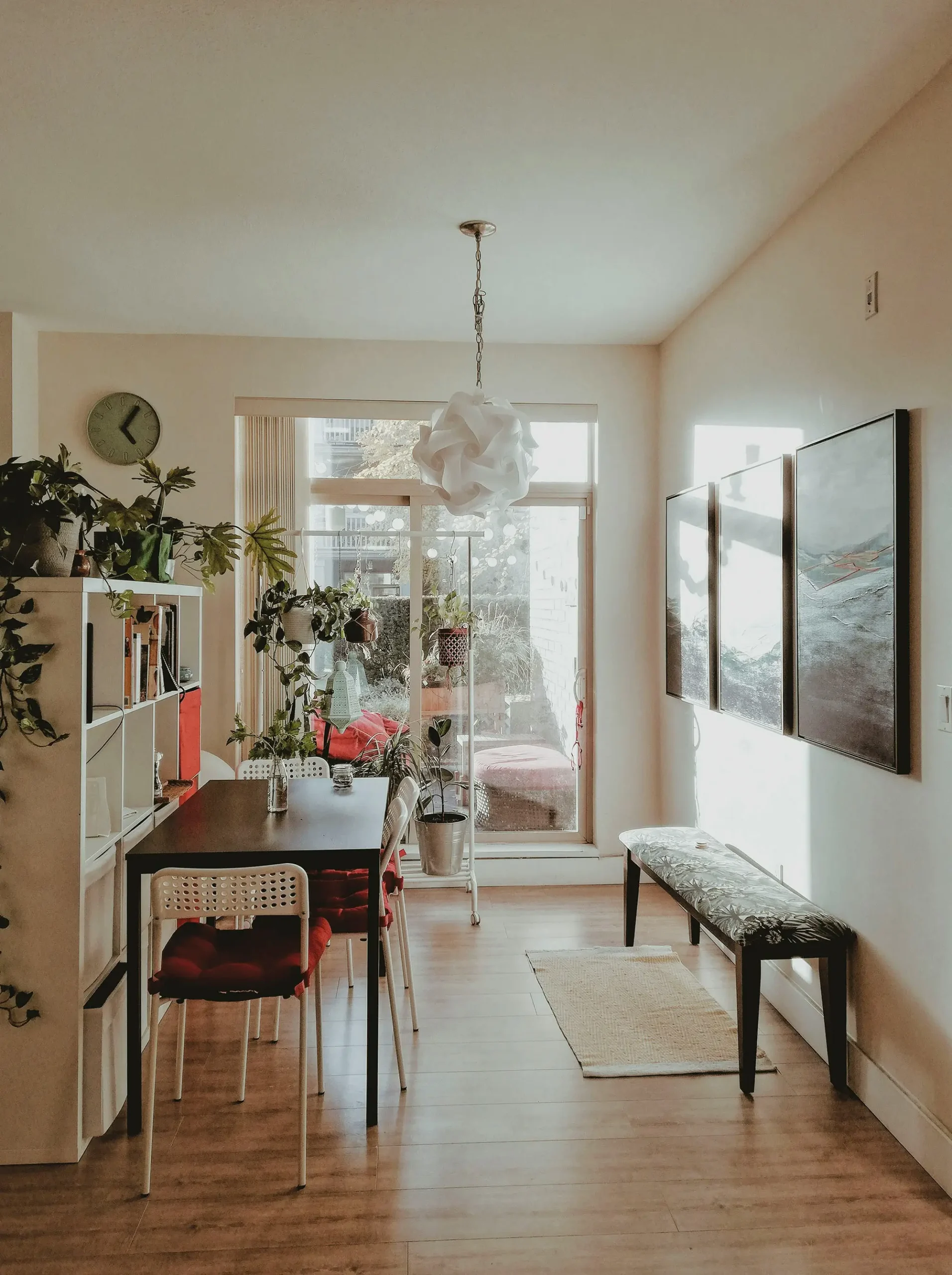 Comedor moderno y acogedor con mesa de madera oscura, sillas blancas, cojines rojos, estanterías con plantas, cuadros decorativos y gran entrada de luz natural desde una puerta de cristal
