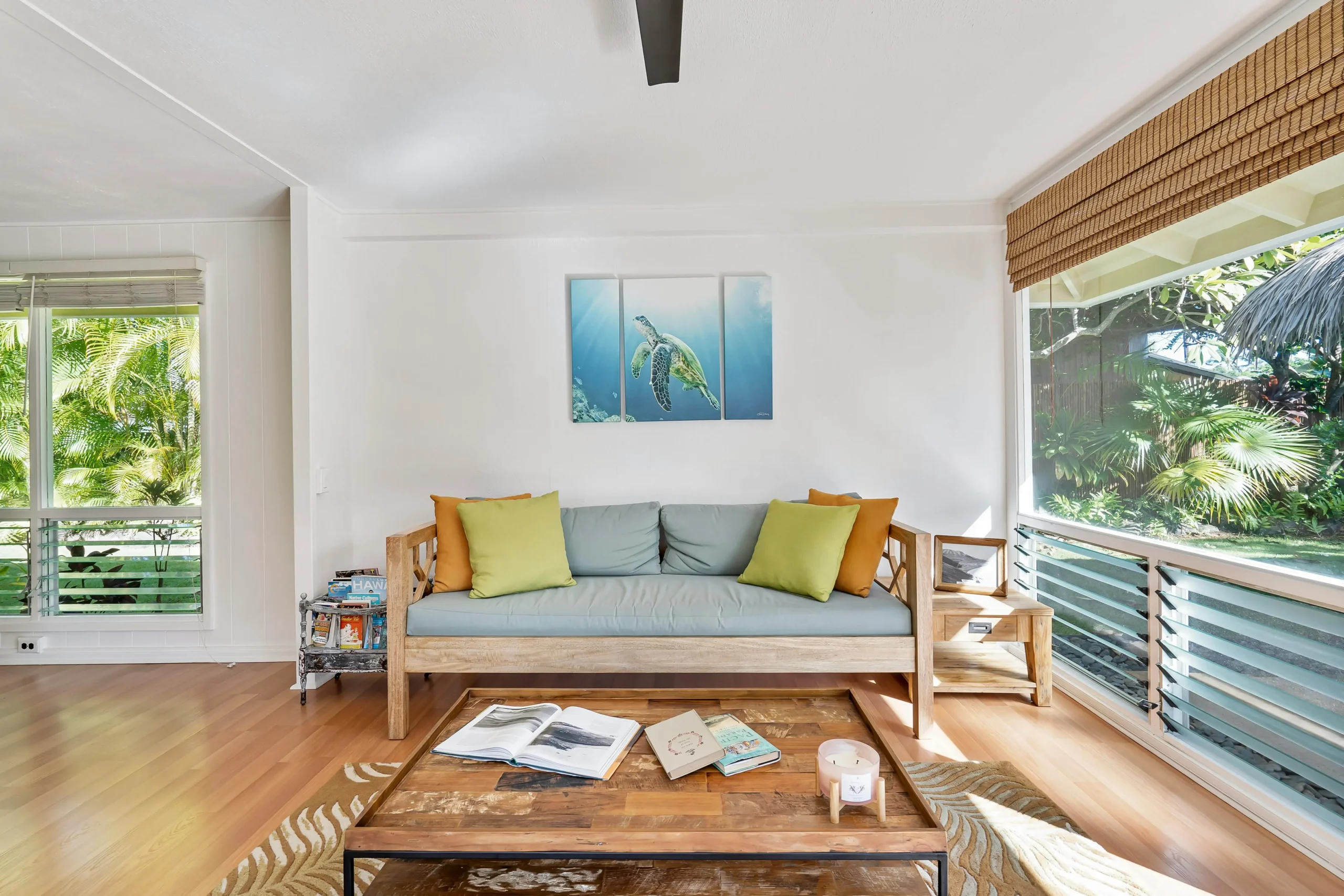 Acogedor salón con sofá de madera clara y cojines en tonos verdes y mostaza, mesa de centro de madera rústica con revistas y velas, grandes ventanales con vistas al jardín tropical y decoración con un cuadro de tortuga marina en la pared