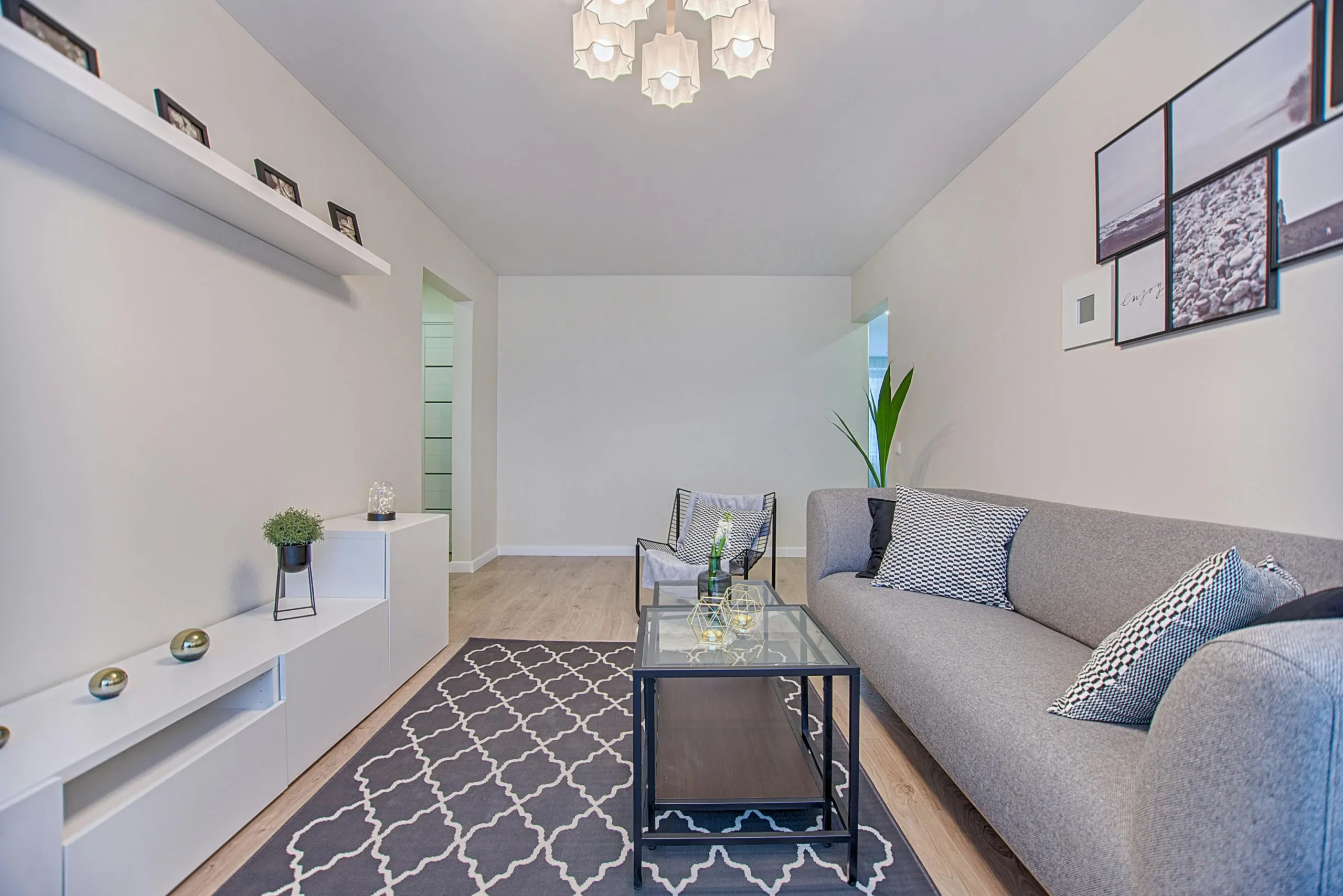 Salón contemporáneo con sofá gris, cojines en blanco y negro, mesa de centro de cristal con estructura metálica, alfombra geométrica, mueble bajo blanco con decoración minimalista, plantas y cuadros en la pared, en un espacio luminoso y ordenado