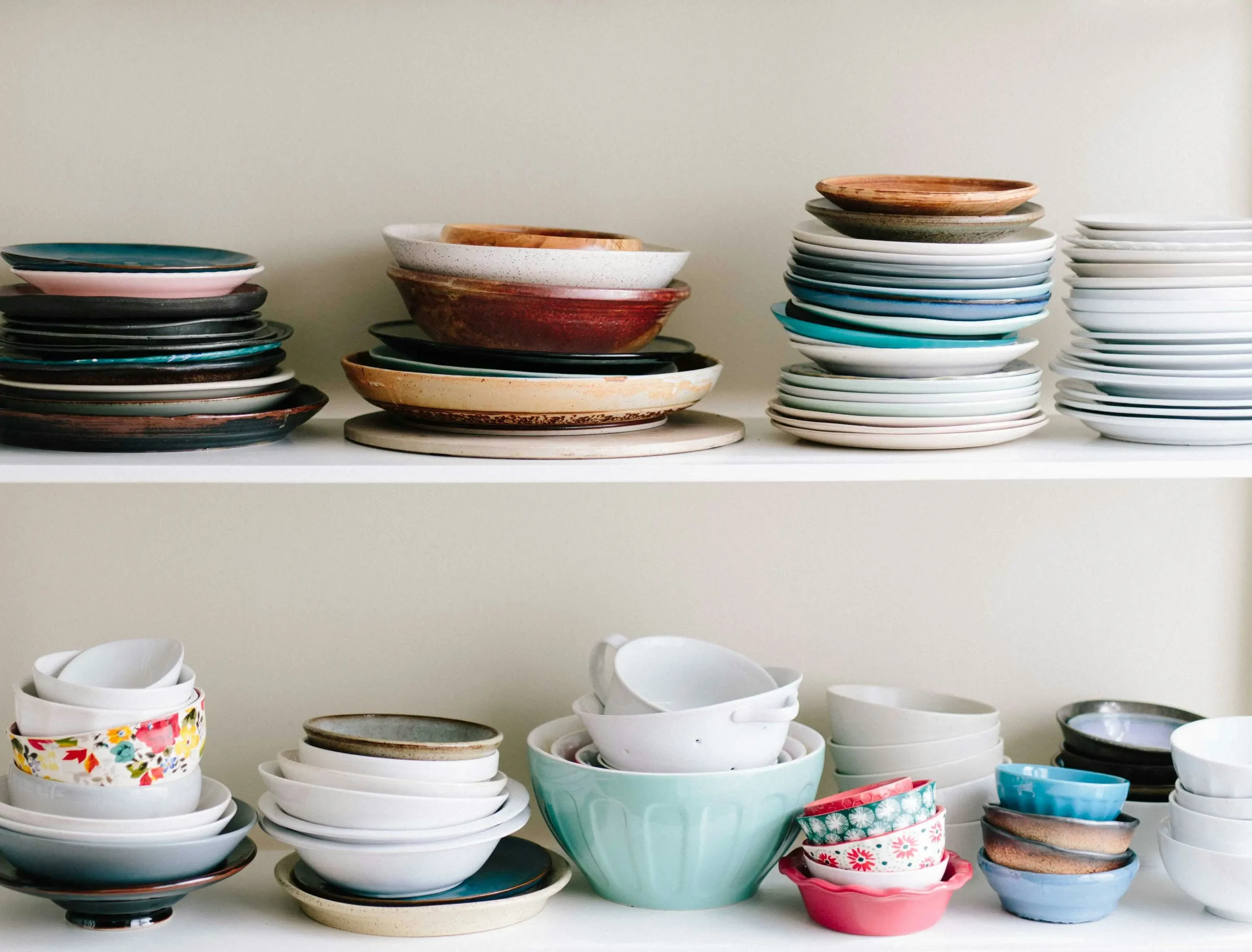 Platos y cuencos de distintos tamaños, colores y estilos apilados de forma ordenada en una estantería blanca de cocina, mostrando una colección variada de vajilla cerámica.