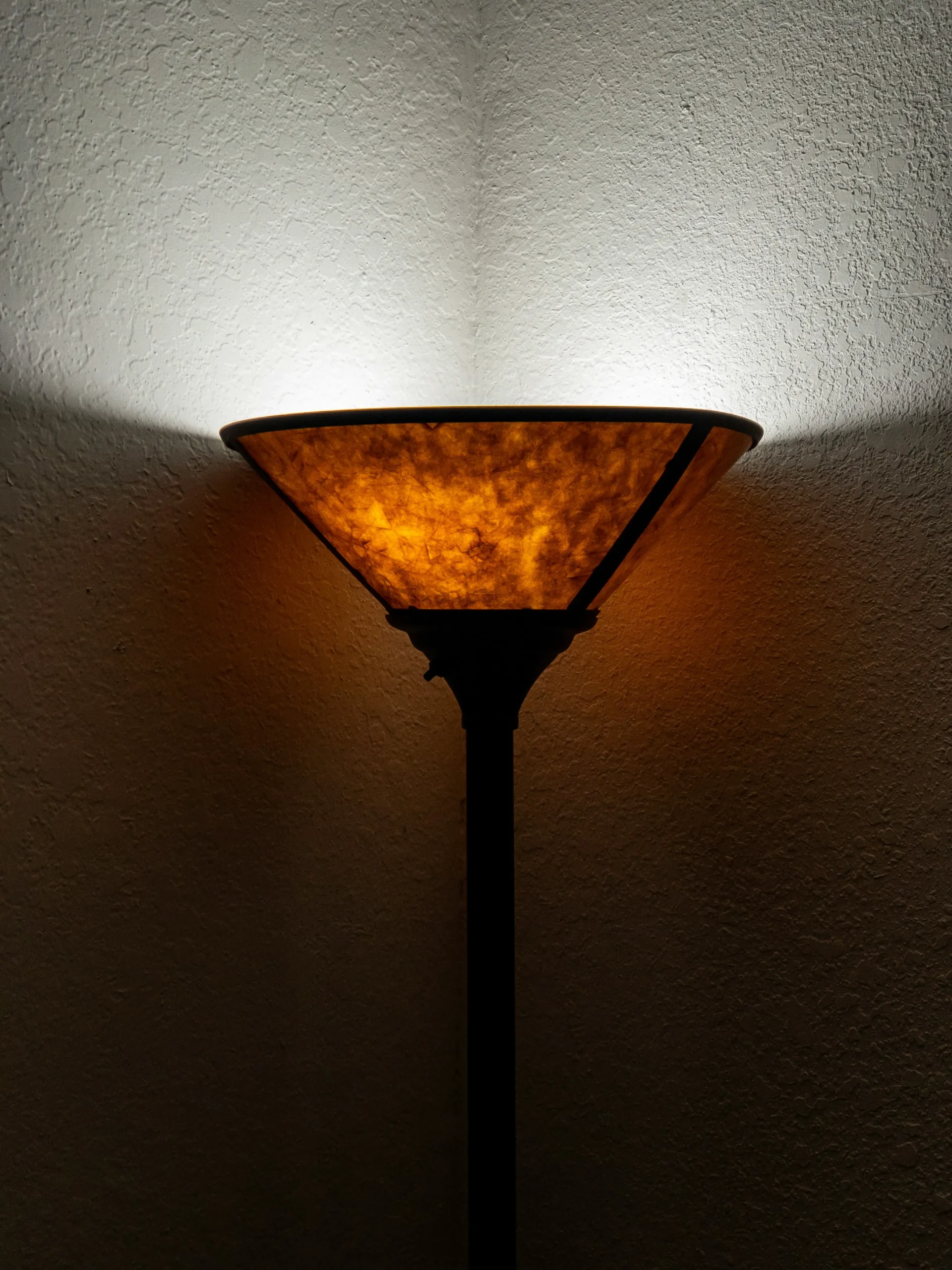 Standing lamp with amber shade lit in the corner of a textured wall, casting warm light and shadows